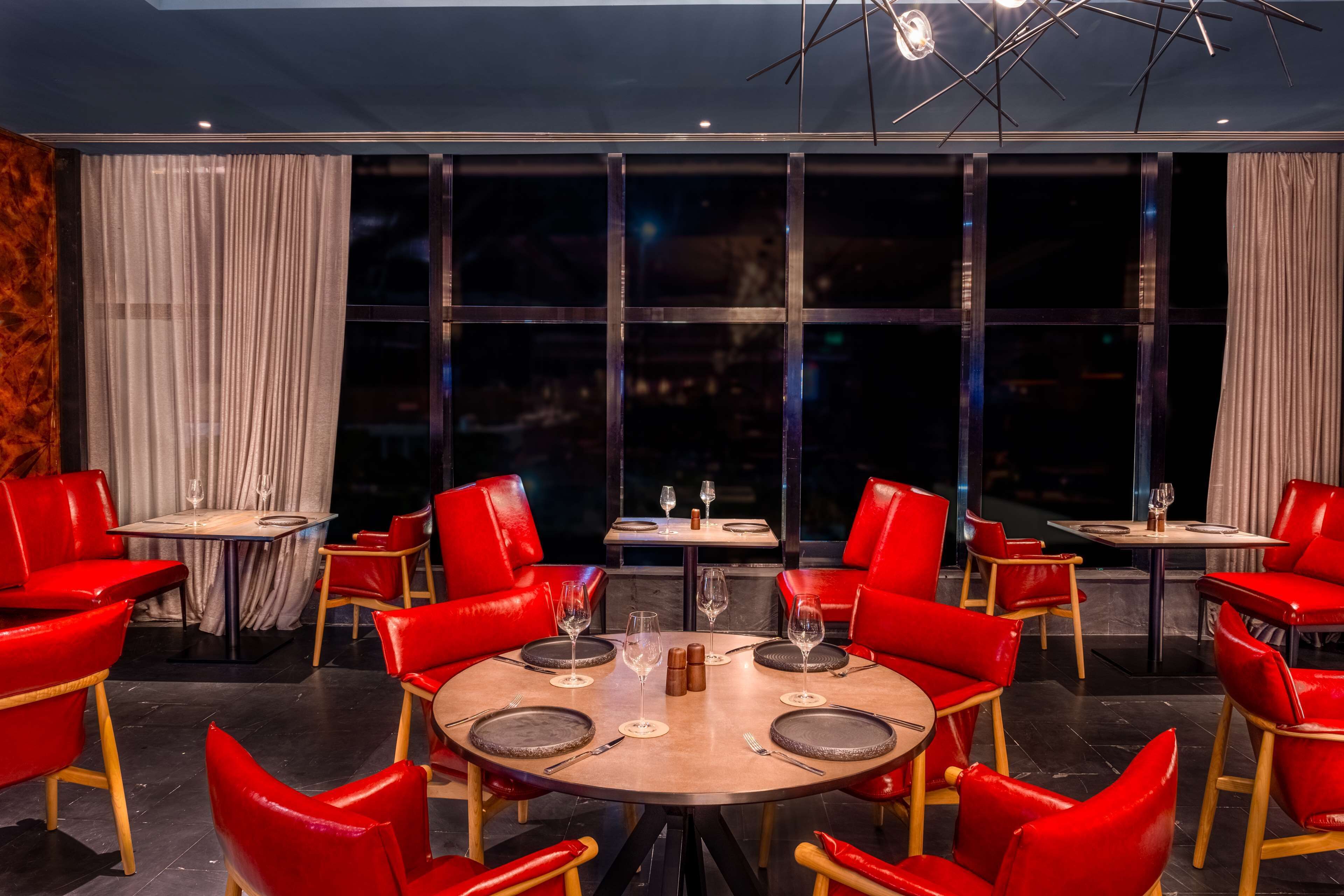A restaurant with round tables and red chairs, large windows, and a view of the city at night.