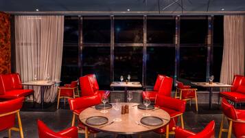 A restaurant with round tables and red chairs, large windows, and a view of the city at night.