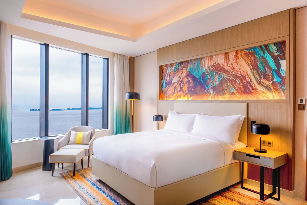 A hotel room with a large bed, a chair, a nightstand, a lamp, and a large window with a view of the water.