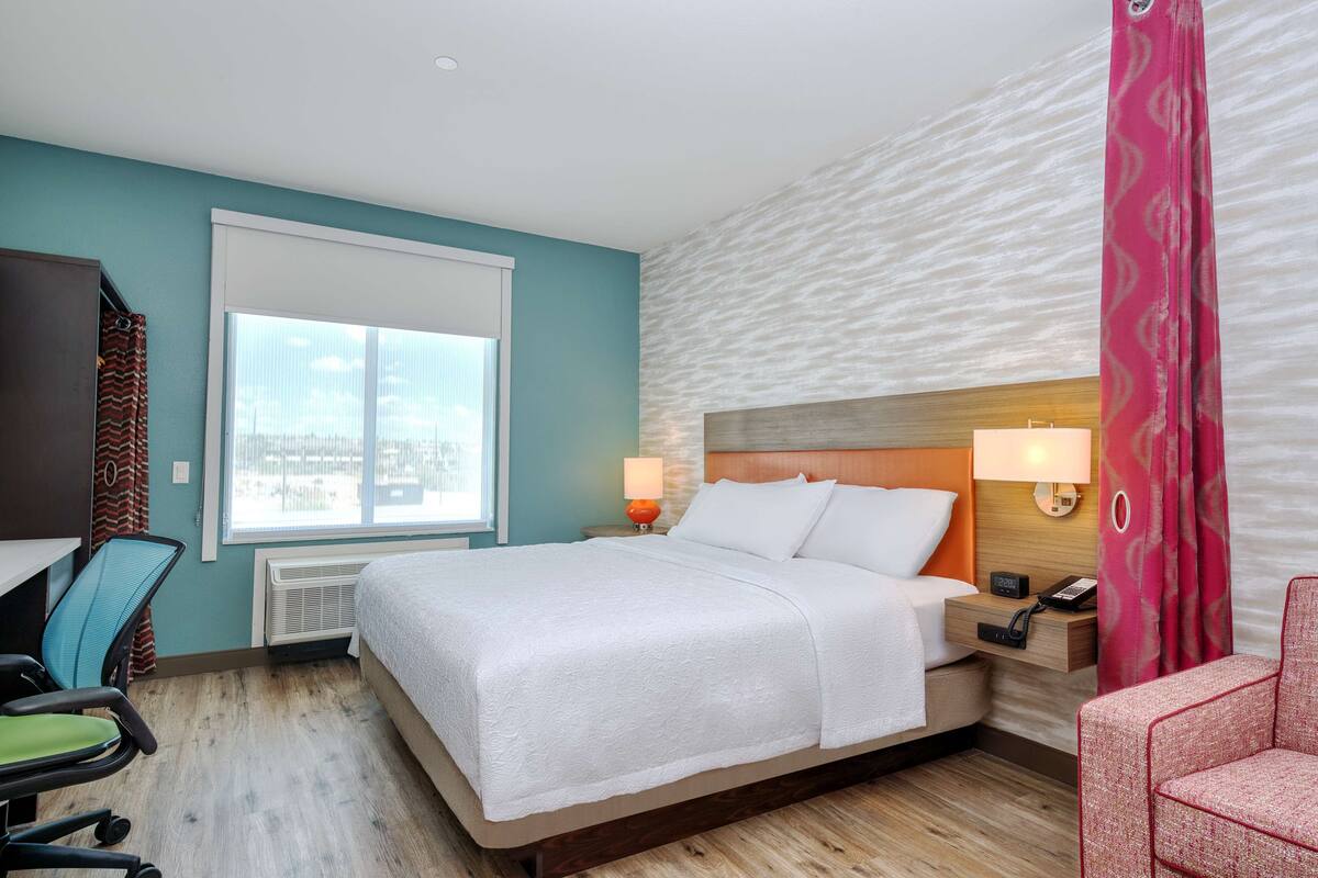 Quarto de hotel com cama, escrivaninha, cadeira e janela com cortinas.