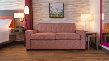 A hotel room with a wooden floor, a red sofa, a bed with white bedding, two bedside lamps, and a framed picture of hot air balloons on the wall.