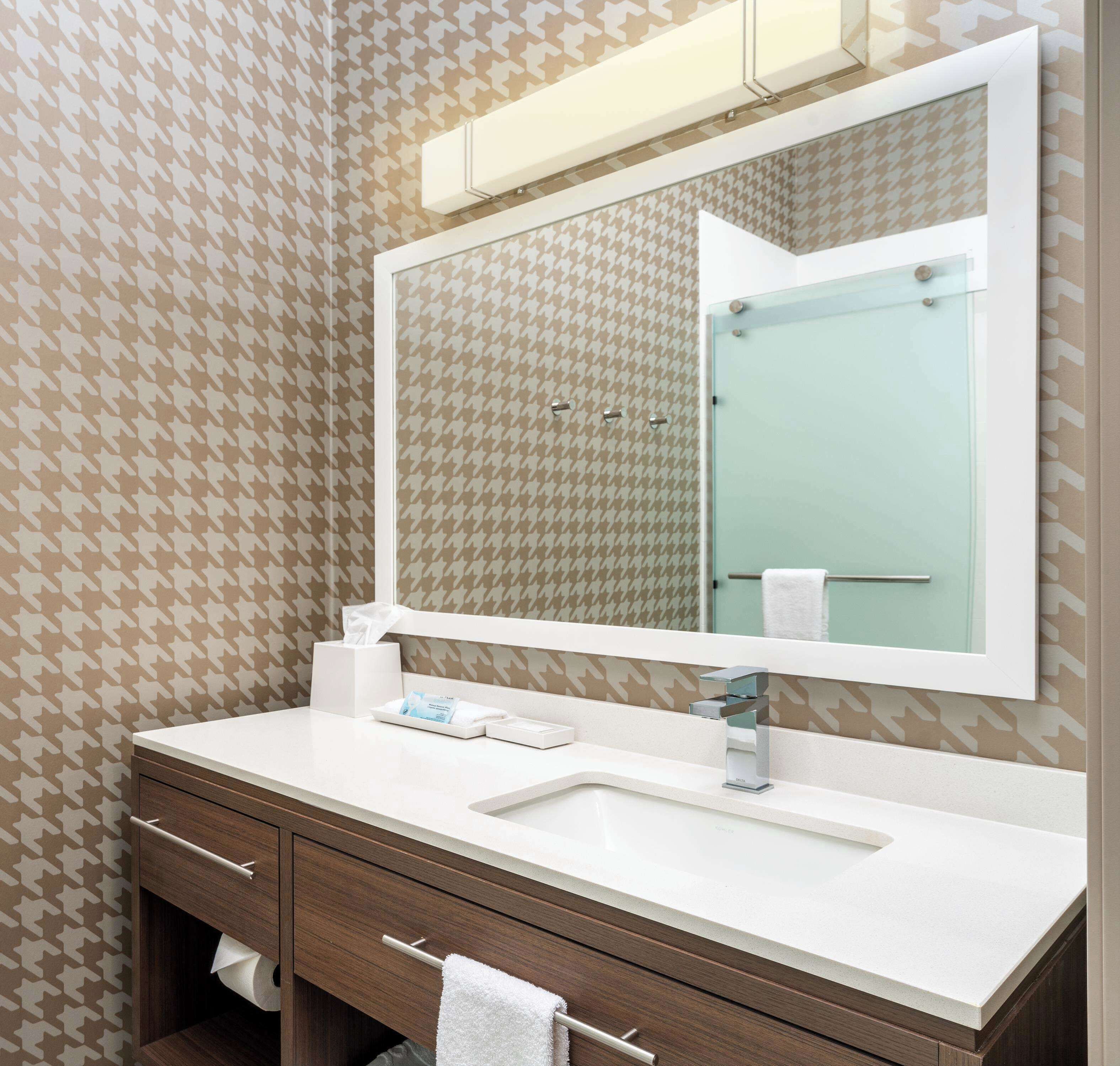 A bathroom with a houndstooth patterned wallpaper, a white sink, a mirror, and wooden cabinetry.