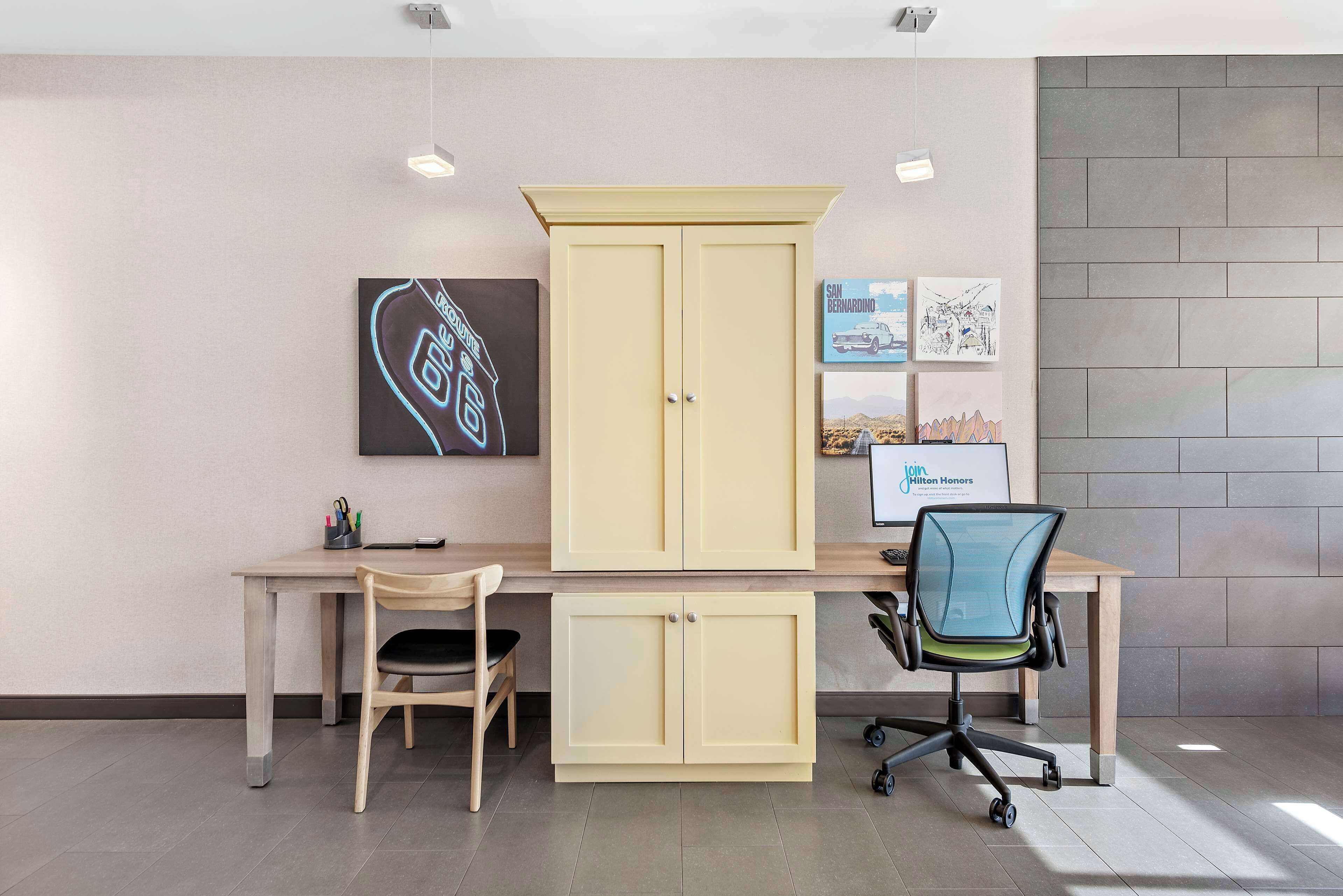 A modern office space with a wooden desk, two chairs, a cream-colored cabinet, and artwork on the wall.
