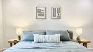 Cama bem arrumada com colcha azul clara e duas almofadas, com dois mesinhas de cabeceira com luminárias e pequenas plantas.