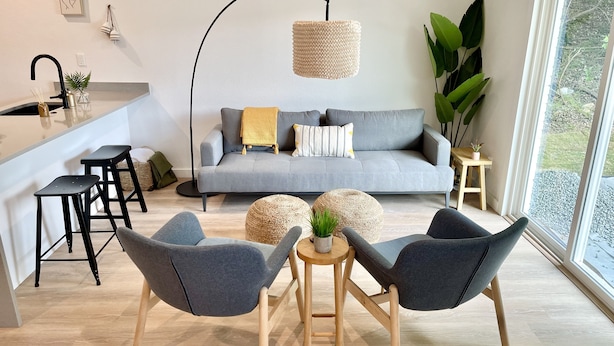Uma sala de estar moderna com um sofá cinzento, duas poltronas cinzentas, uma mesa de centro e uma área de cozinha com uma torneira preta e banquetas.