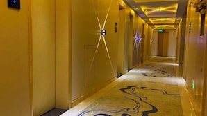 A hotel hallway with a patterned carpet, wall-mounted light fixtures, and a ceiling with recessed lighting. - Wuyi E-Sports Hotel (Chenzhou)