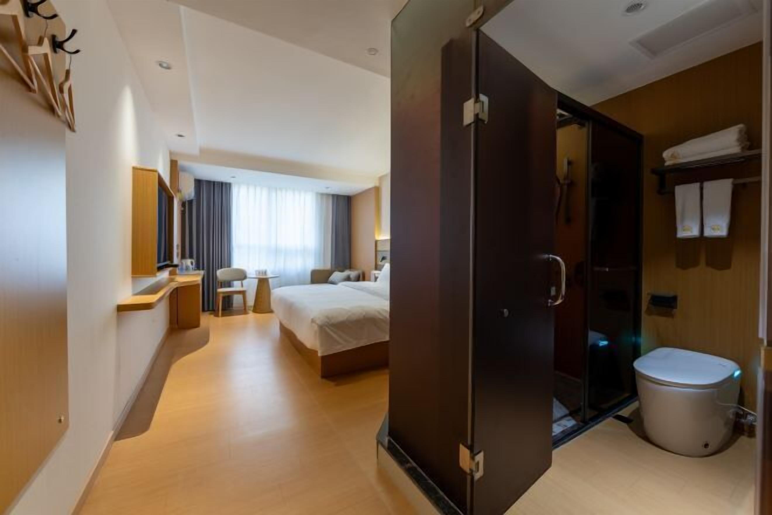 A hotel room with a bed, a desk, a chair, a mirror, a window with curtains, a bathroom with a toilet, and a shower area.