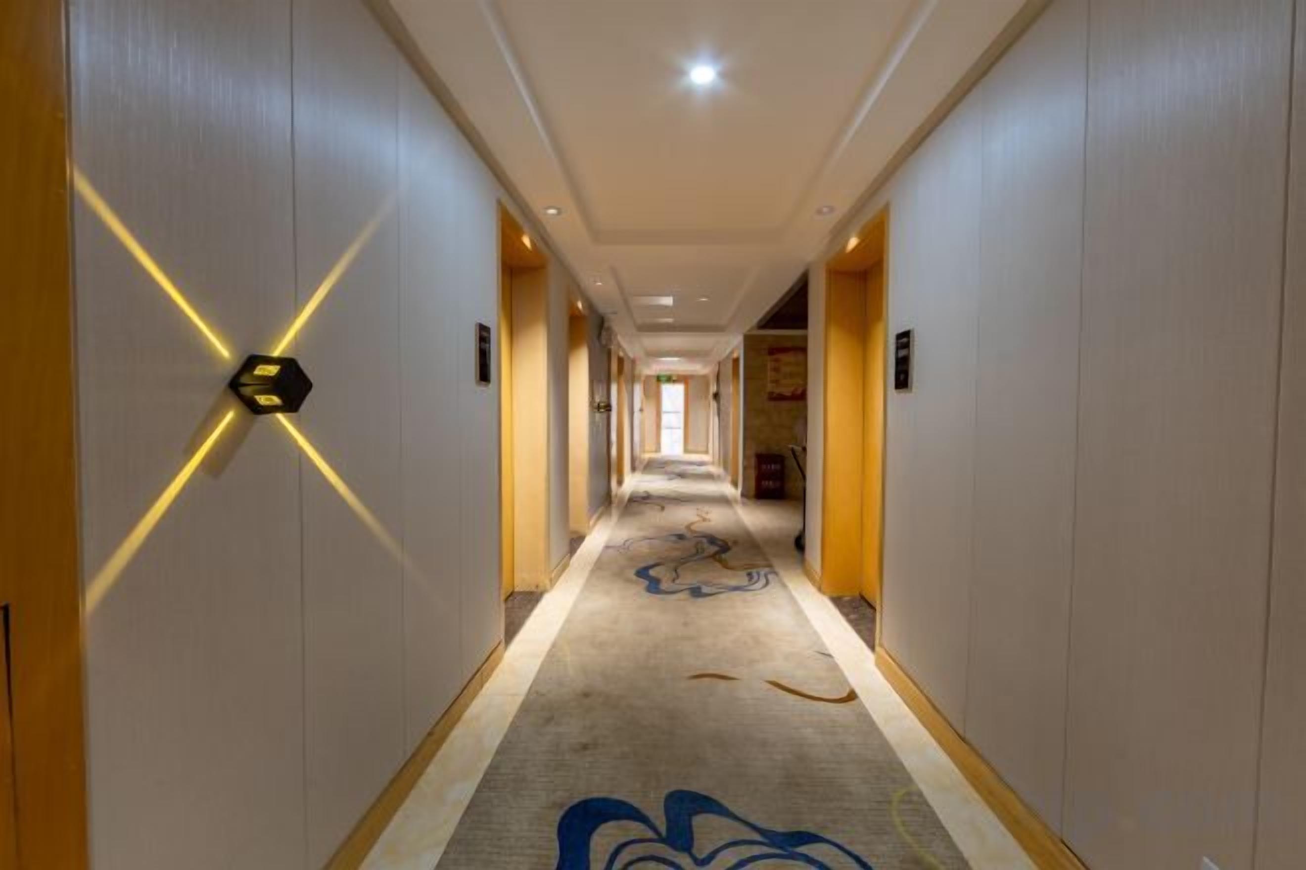 A hotel corridor with a carpeted floor, a light-colored wall with a geometric design, and a door at the end.