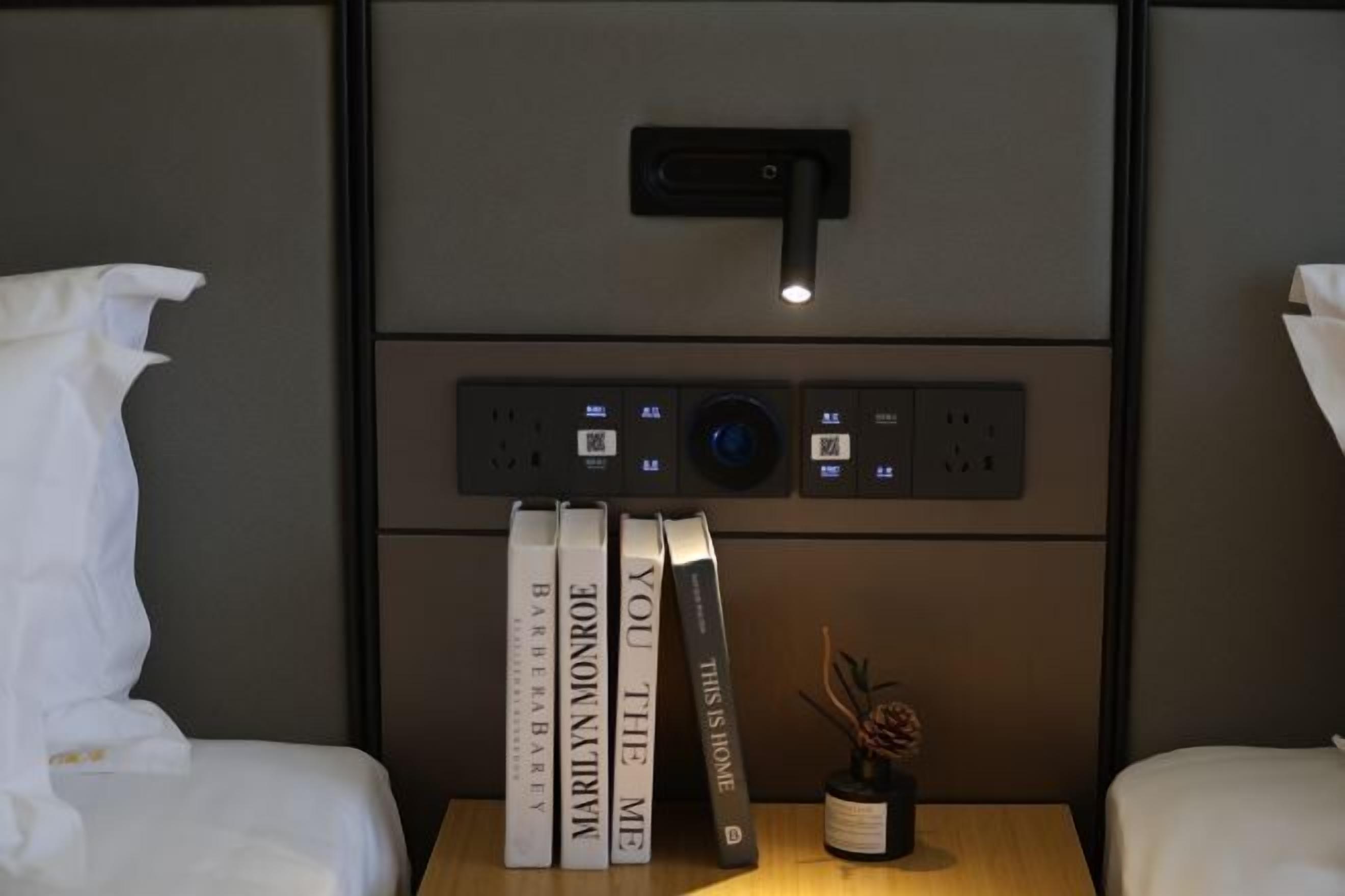 A hotel room with a bedside table, a lamp, and a wall-mounted control panel.