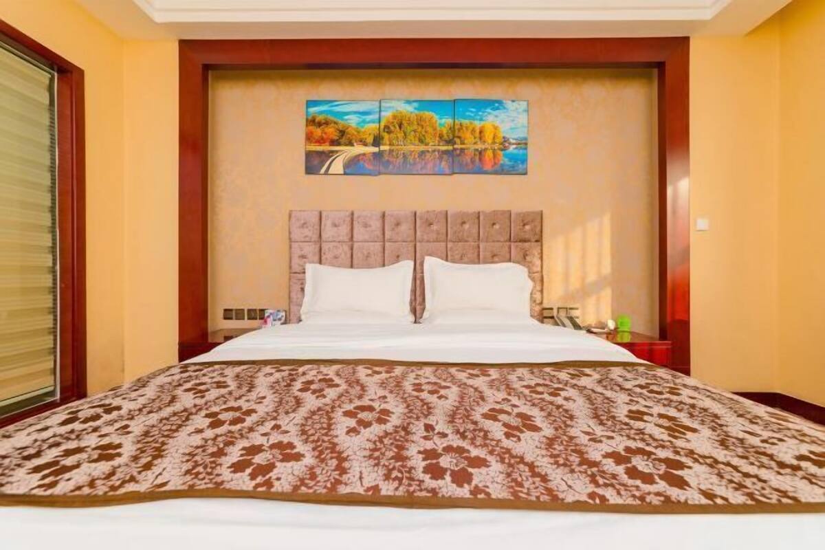 Une chambre d’hôtel avec un grand lit, une tête de lit ornée d’un tableau représentant un paysage pittoresque et une fenêtre avec des stores.