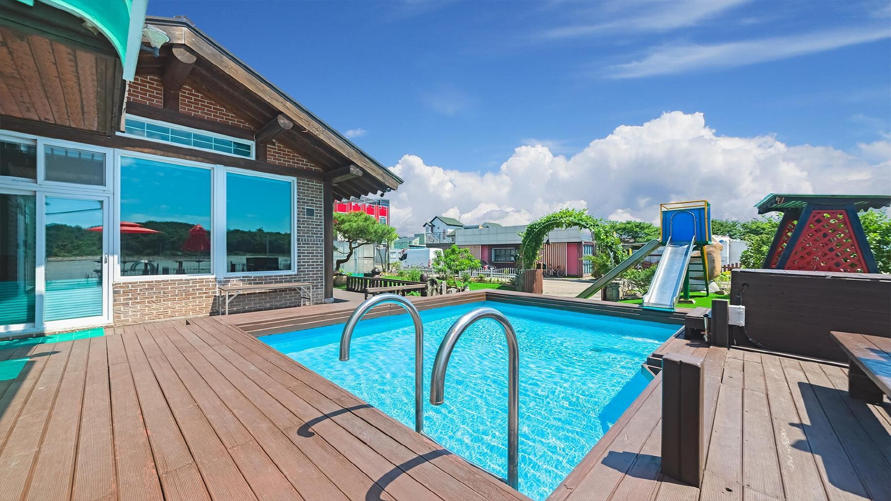 Un área de piscina con terraza de madera, un edificio con ventanas grandes y un parque infantil con toboganes.