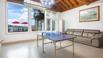 Una habitación con mesa de ping pong, sofá de cuero, un ventanal con vista a un muelle y sombrillas, y un cuadro con un paisaje.