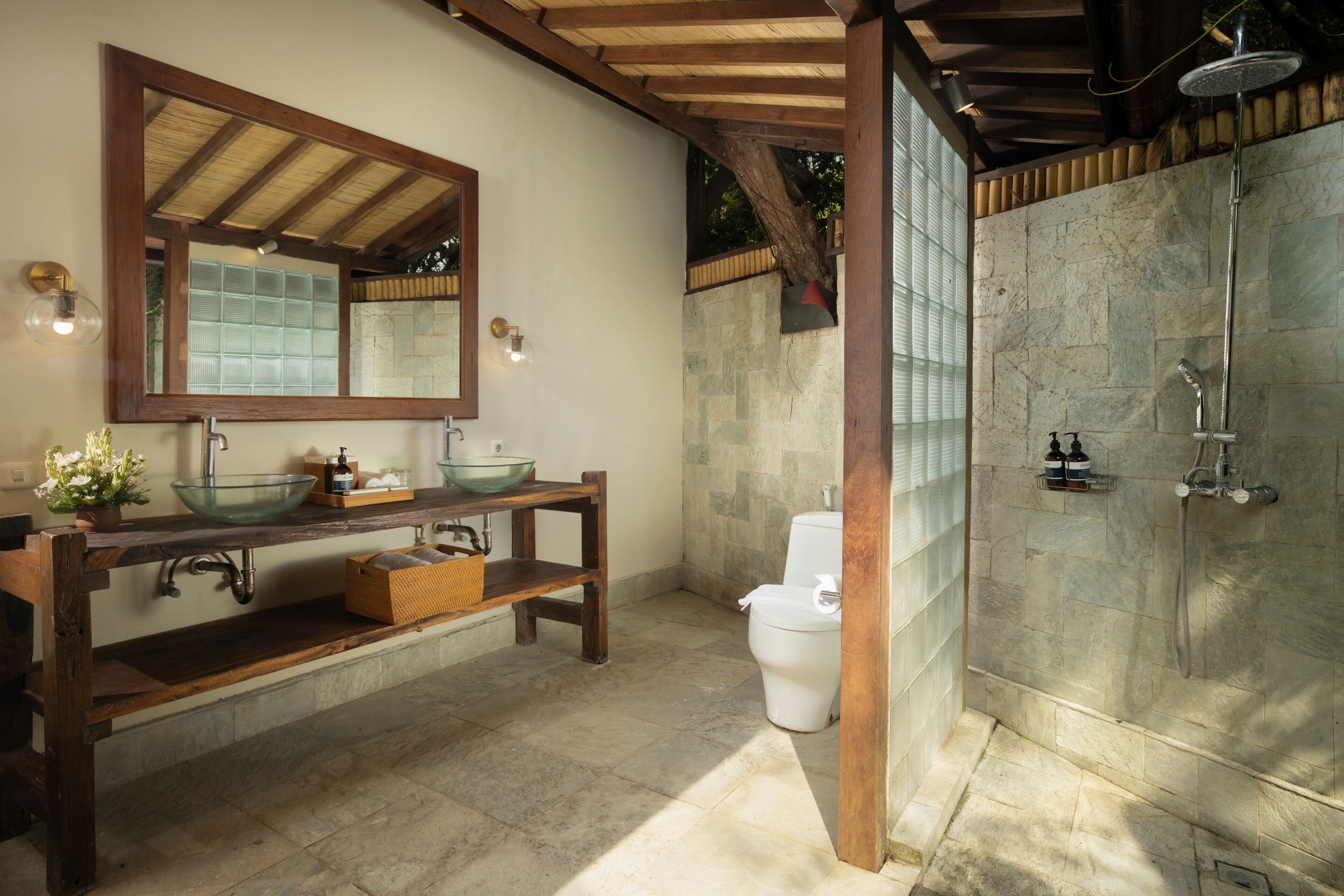 Une salle de bain avec un meuble-lavabo en bois, un lavabo en forme de bol en verre, des toilettes blanches et une douche à l’italienne avec des parois en pierre.