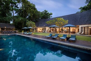 Ein Swimmingpool mit Liegestühlen und Sonnenschirmen, umgeben von einem Gebäude mit überdachter Terrasse und Außenbestuhlung.