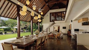 A spacious dining area with a long wooden table set for six, surrounded by wicker chairs. A kitchen area with a large island and bar stools is visible to the right. - Villa Tirtadari by Nakula (Kerobokan)