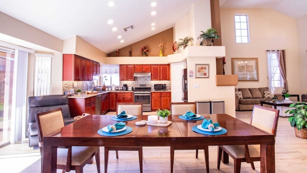 A spacious living area with a dining table set for four, a kitchen with red cabinets, and a large window providing natural light. - Private Vegas Villa with Pool & Spa Perfect for a European Escape (North Las Vegas)