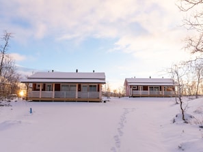 Two cabins with a snow-covered ground, a clear sky, and a sunset in the background.