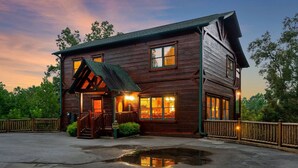 Une cabane en rondins à deux étages, dotée d’une véranda, de grandes fenêtres et d’une entrée bien éclairée.