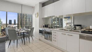 Une cuisine moderne avec des meubles blancs, un coin repas avec des chaises grises et une grande fenêtre offrant une vue sur la ville.