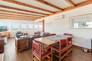 Une pièce chaleureuse avec un plafond en bois, une grande fenêtre donnant sur la mer, une table à manger avec des chaises et un canapé agrémenté de coussins à motifs.