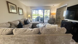 A living room with a beige sofa, a coffee table, a TV, and a sliding glass door leading to an outdoor patio.