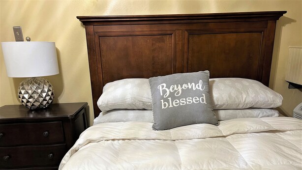 Ein ordentlich bezogenes Bett mit einem Kissen, auf dem 'Beyond blessed' steht, und eine geometrisch gestaltete Lampe auf einem Nachttisch.