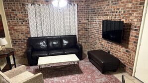Ein Wohnzimmer mit Backsteinwand, einer schwarzen Ledersofa, einem Couchtisch, einem gemusterten Sessel, einem rot-weißen Teppich und einem wandmontierten Flachbildfernseher.