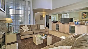 Un salón amplio con un conjunto de sofás, una mesa de centro y un televisor. Se observa una zona de cocina con armarios y un televisor.