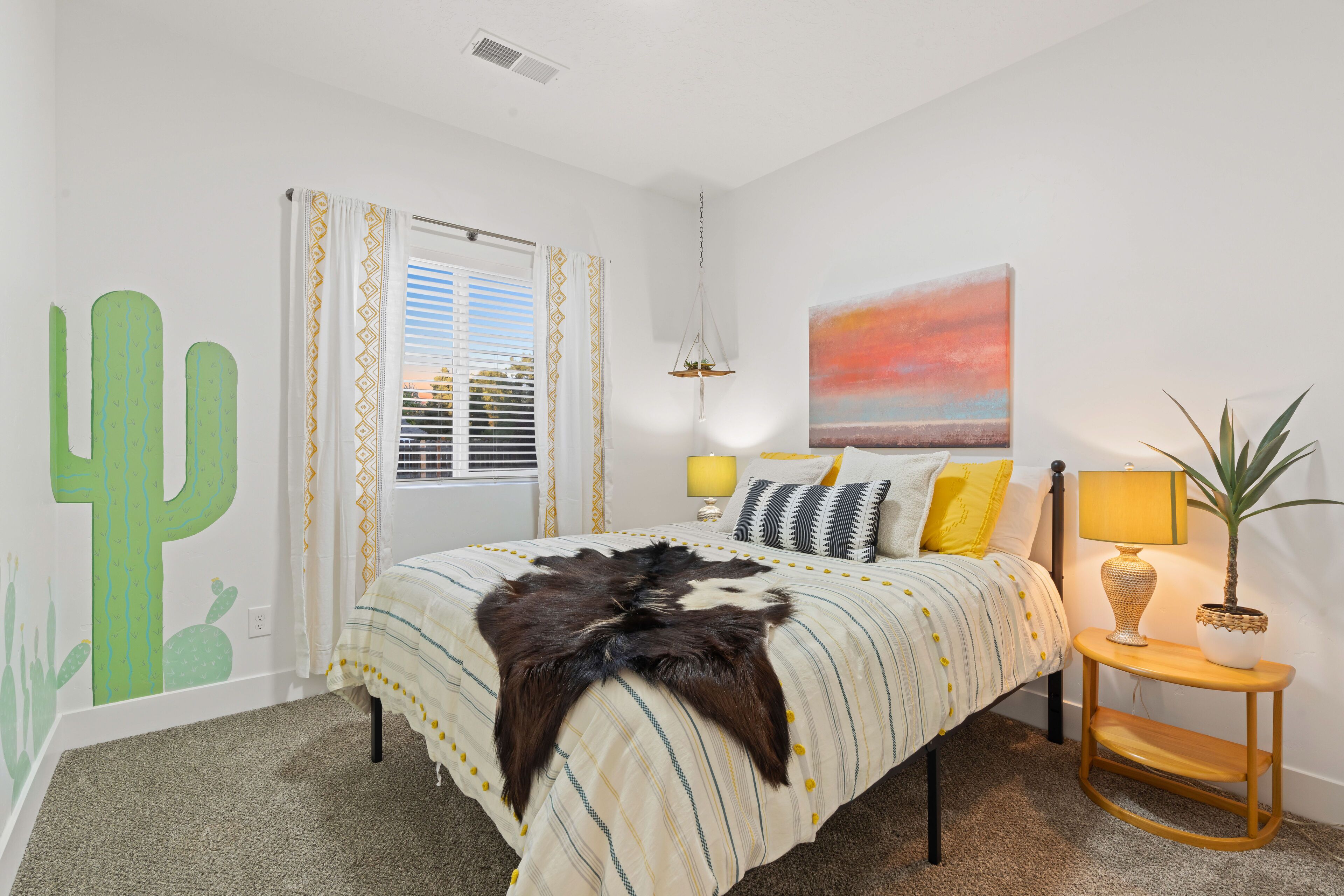 A bedroom with a bed, a nightstand, a lamp, a cactus wall decal, a window with curtains, and a painting on the wall.