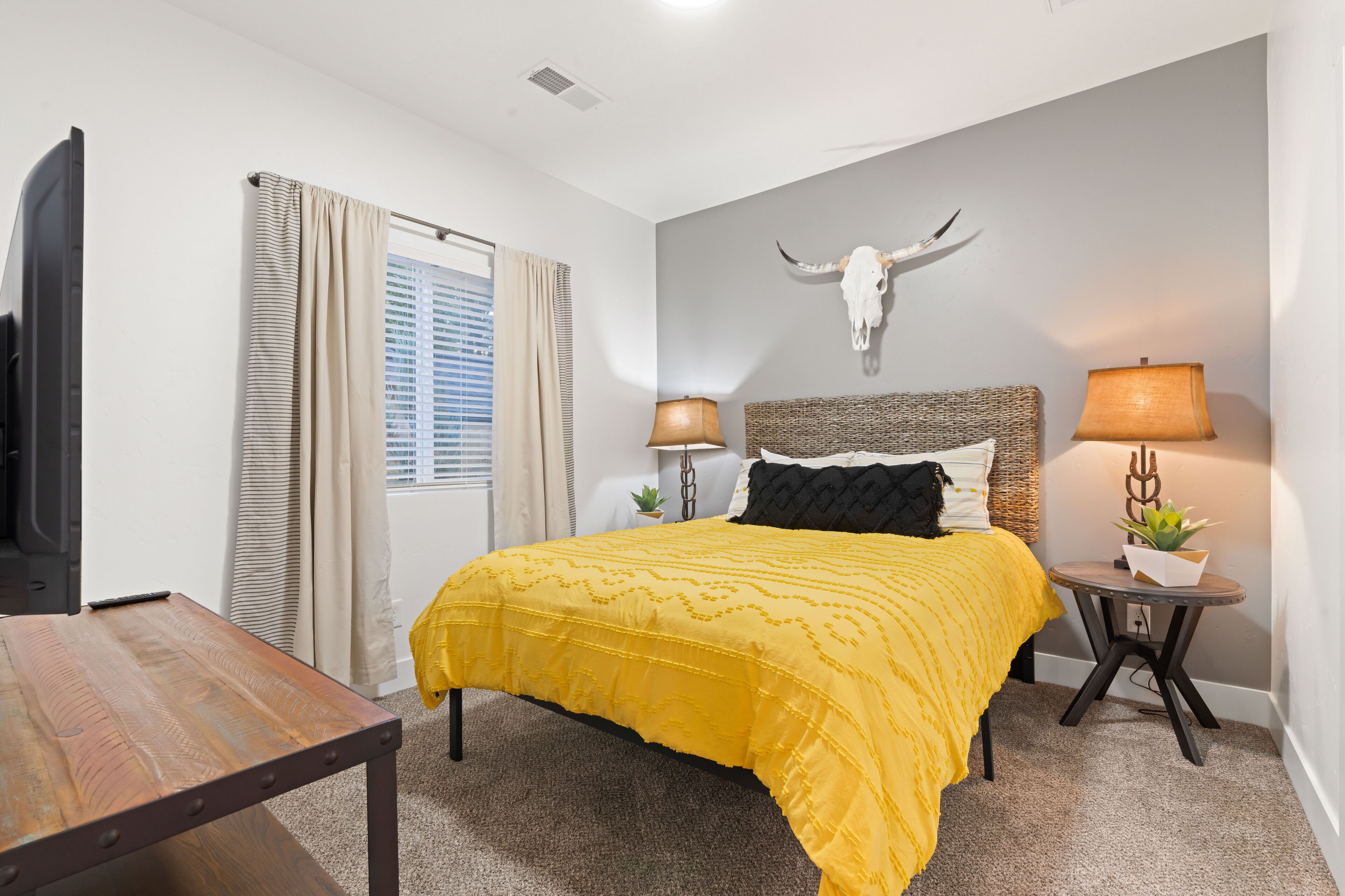 A bedroom with a bed, bedside lamps, a mounted cow skull, and a window with curtains.