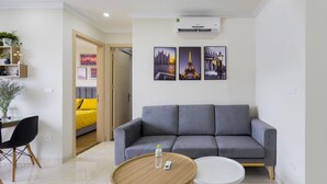 A modern living room with a grey sofa, a round wooden coffee table, and a white side table. There are framed pictures on the wall, a small shelf with plants, and a door leading to another room.