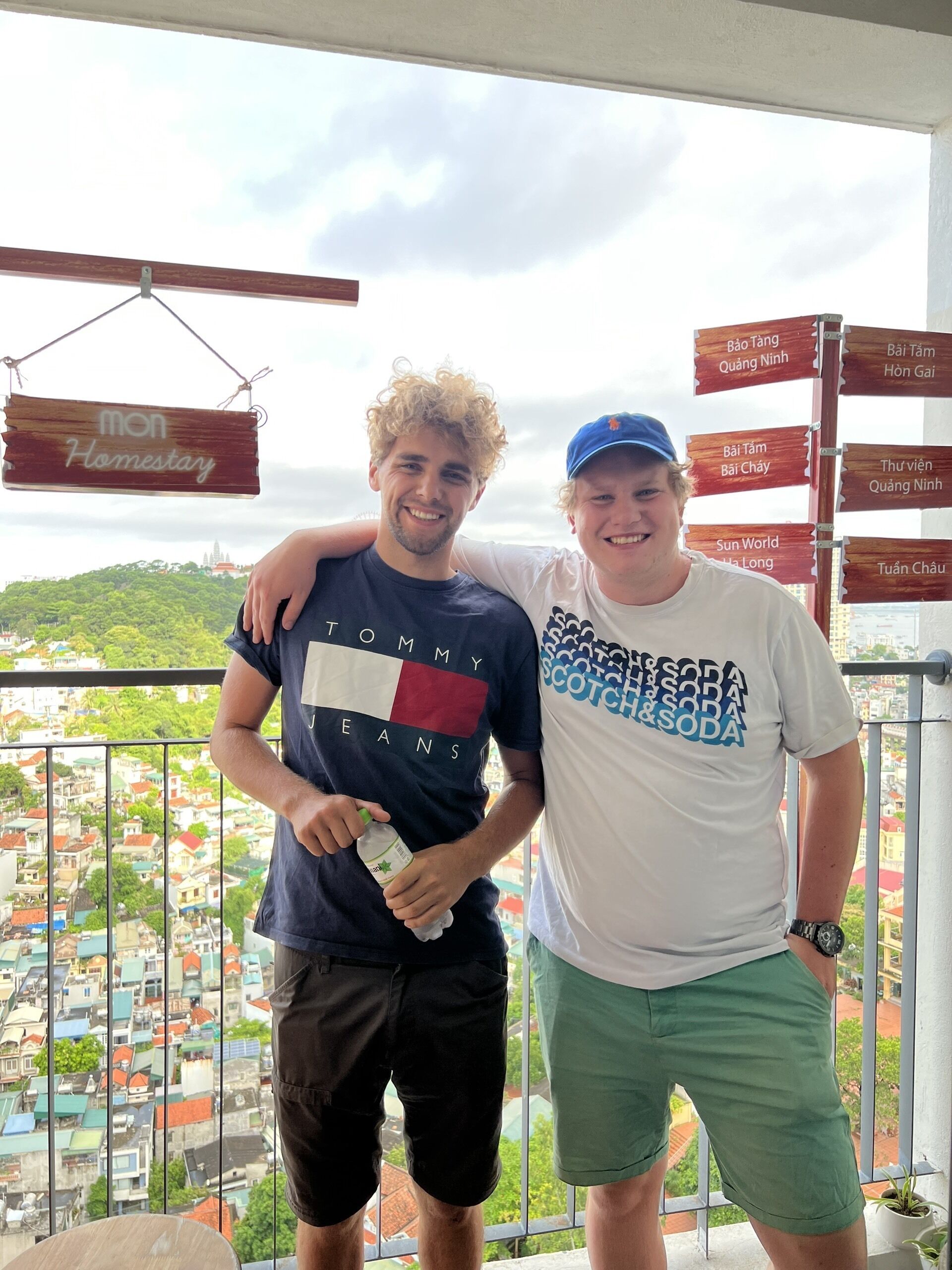 Two people standing on a balcony with a city view, a sign reading "Homestay", and multiple directional signs.