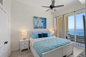 Een slaapkamer met een bed, nachtkastjes, een plafondventilator, een schilderij en uitzicht op de oceaan door een schuifdeur.