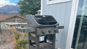 Una barbacoa negra d'exterior amb una llar de foc integrada, situada en un patio de formigó amb vista a una casa i muntanyes al fons.
