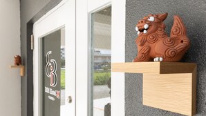 A modern building entrance with a glass door, a wooden shelf with a decorative lion figure, and a sign with a logo and text. - BIBI HOTEL MIYAKO KUKOMAE (Miyakojima)