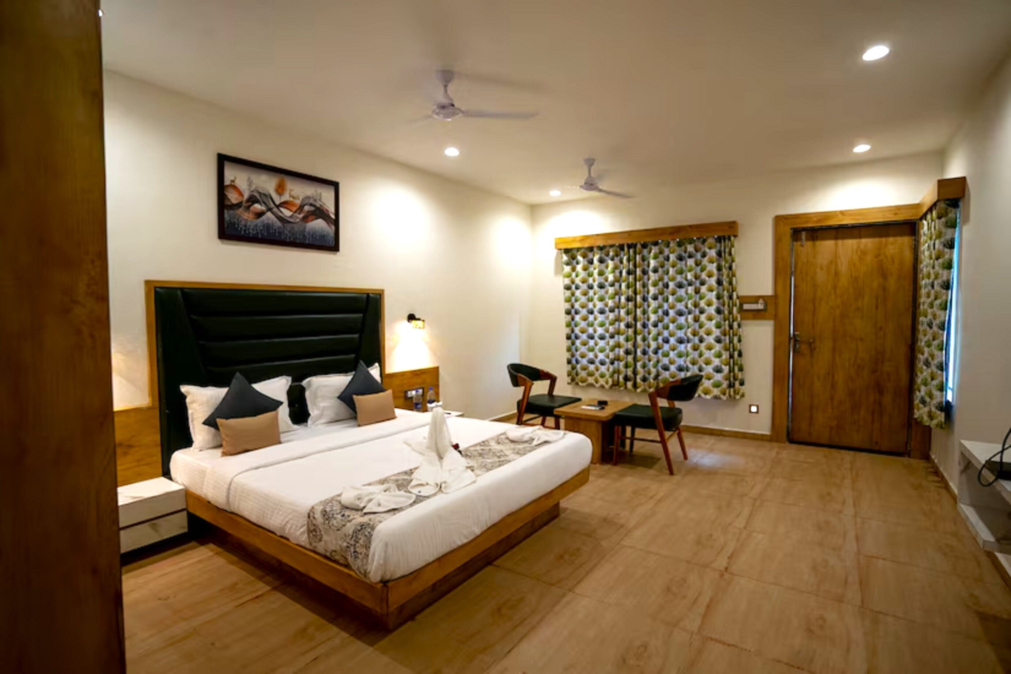 A hotel room with a large bed, a desk, two chairs, a ceiling fan, and a painting on the wall.