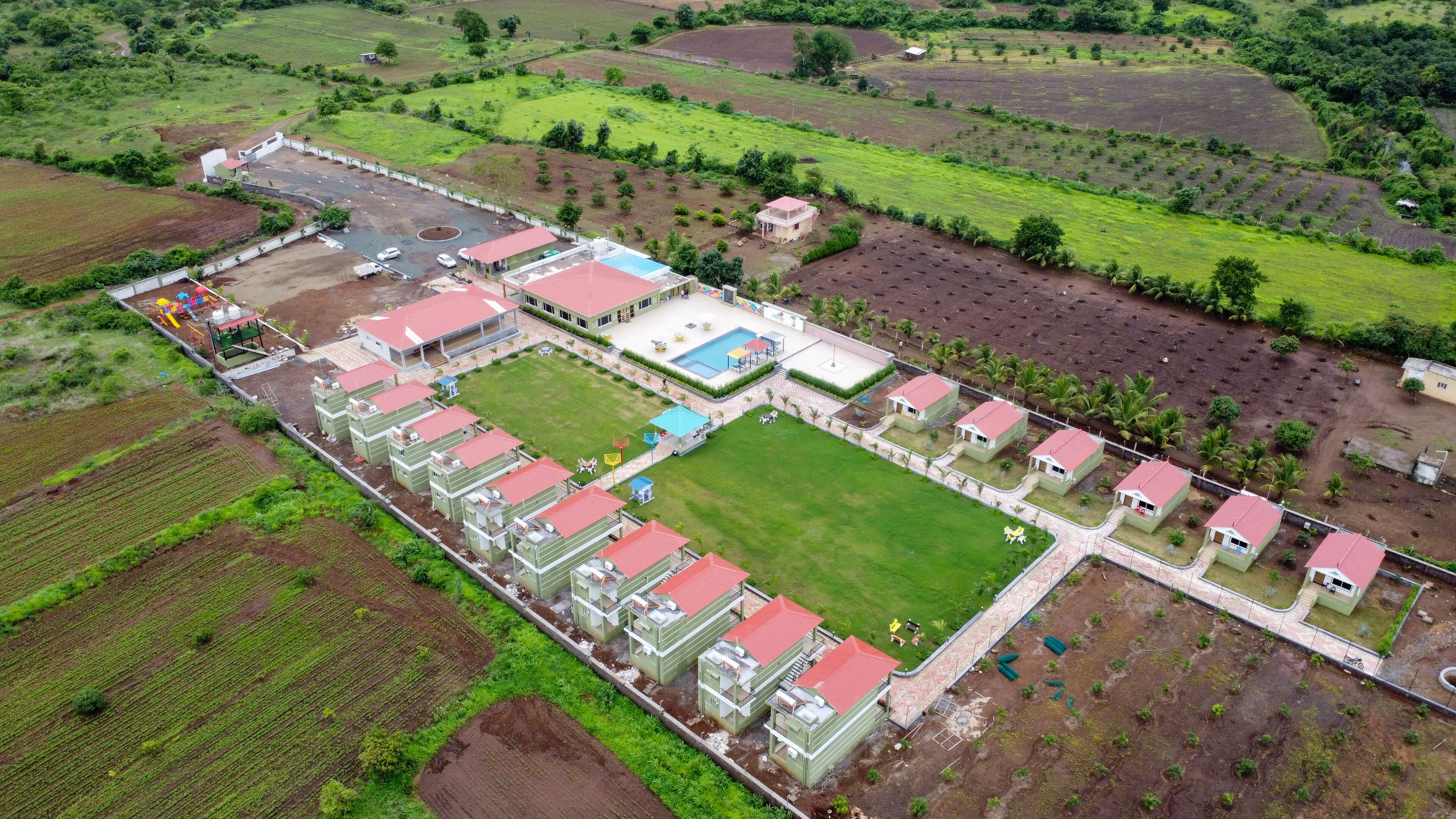 An aerial view of a resort with multiple buildings, a swimming pool, and surrounding green fields.