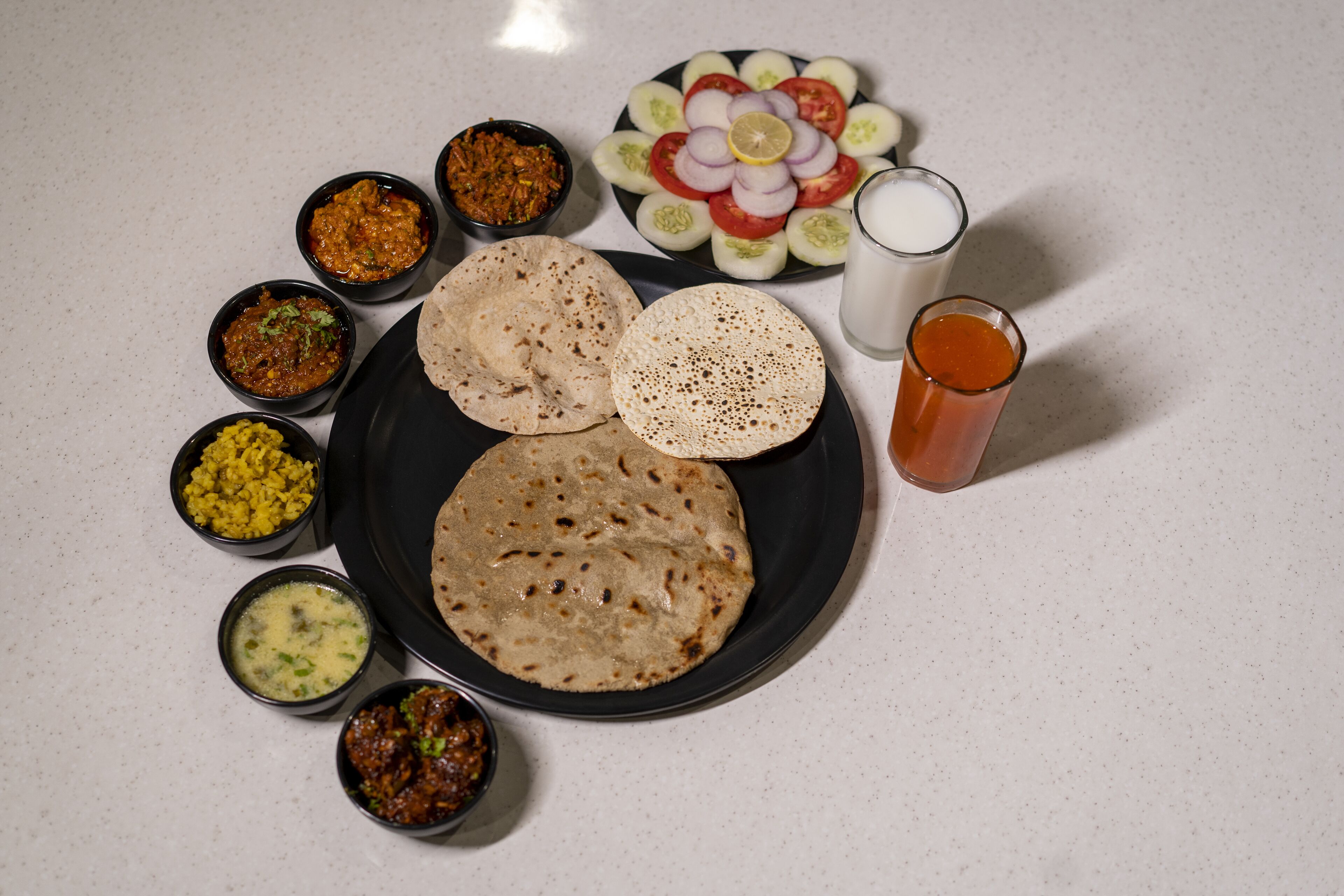 A plate with two flatbreads, a variety of chutneys, sliced vegetables, and a glass of milk.