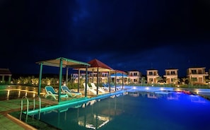 A nighttime swimming pool area with lounge chairs and a covered seating area.