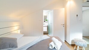 Un dormitorio moderno con cama, mesita de noche, un taburete pequeño y un baño visible a través de una puerta abierta.