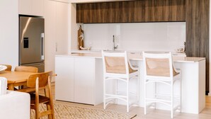 Eine moderne Küche mit weißen Schränken, einem Edelstahlkühlschrank und einer Wand in Holzoptik. Zwei weiße Barhocker mit Geflochtener Sitzfläche sind vorhanden.