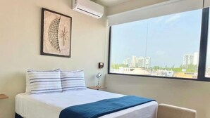 A hotel room with a bed, a chair, a window with a city view, and a wall-mounted air conditioner. - Rooftop Pool Apt for 4 | Near Isla Ferry, Cancun (CancÃºn)