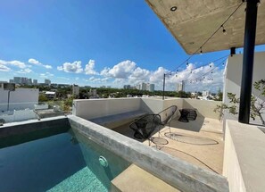 Une piscine sur le toit offrant une vue dégagée sur les gratte-ciel de la ville, un espace détente avec des chaises, et une structure en béton au-dessus.