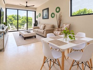 A modern living room with a sofa, ottoman, dining table, and chairs. There is a balcony with a view of greenery.