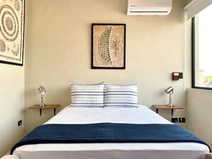 Un lit bien fait, avec un oreiller à rayures blanches et bleues, une couvre-lit bleu foncé et une œuvre d’art représentant un croissant de lune accrochée au mur.