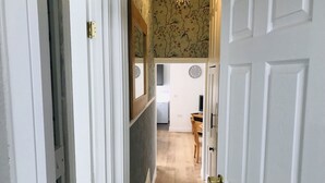 Un couloir mène à une salle à manger dont le sol est décoré de motifs, où l’on aperçoit une horloge au mur et une table en bois.