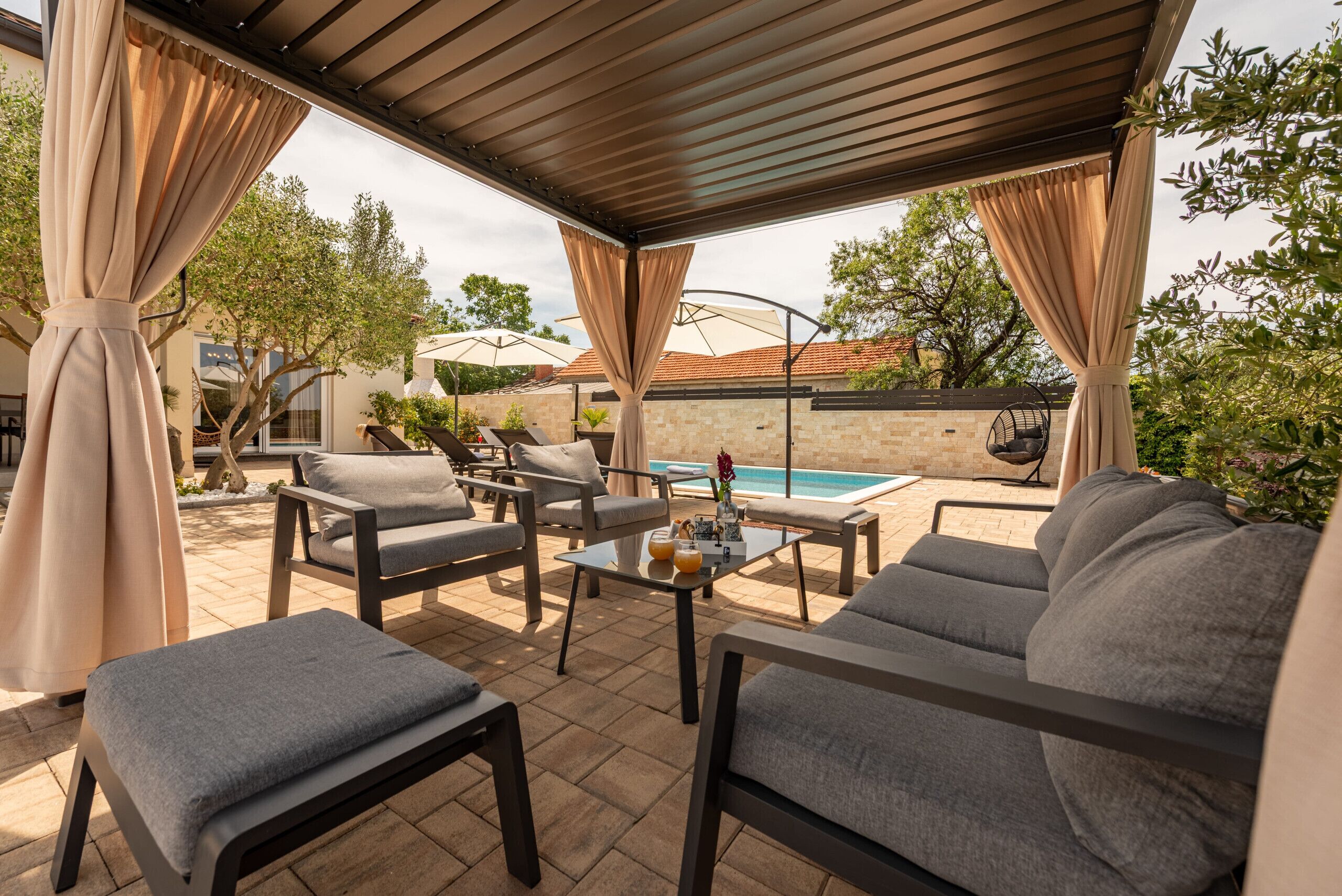 Un espacio al aire libre con patio, asientos acolchados, una mesa de vidrio y una piscina al fondo.