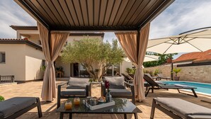Zona relax esterna coperta con poltrone, tavolino con vassoi di snack e bevande, e piscina sullo sfondo.