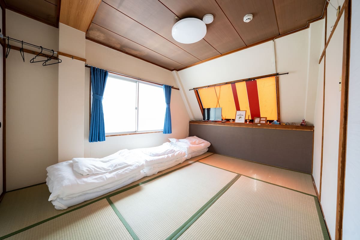 A room with tatami flooring, a bed, a window with blue curtains, and a shelf with various items.
