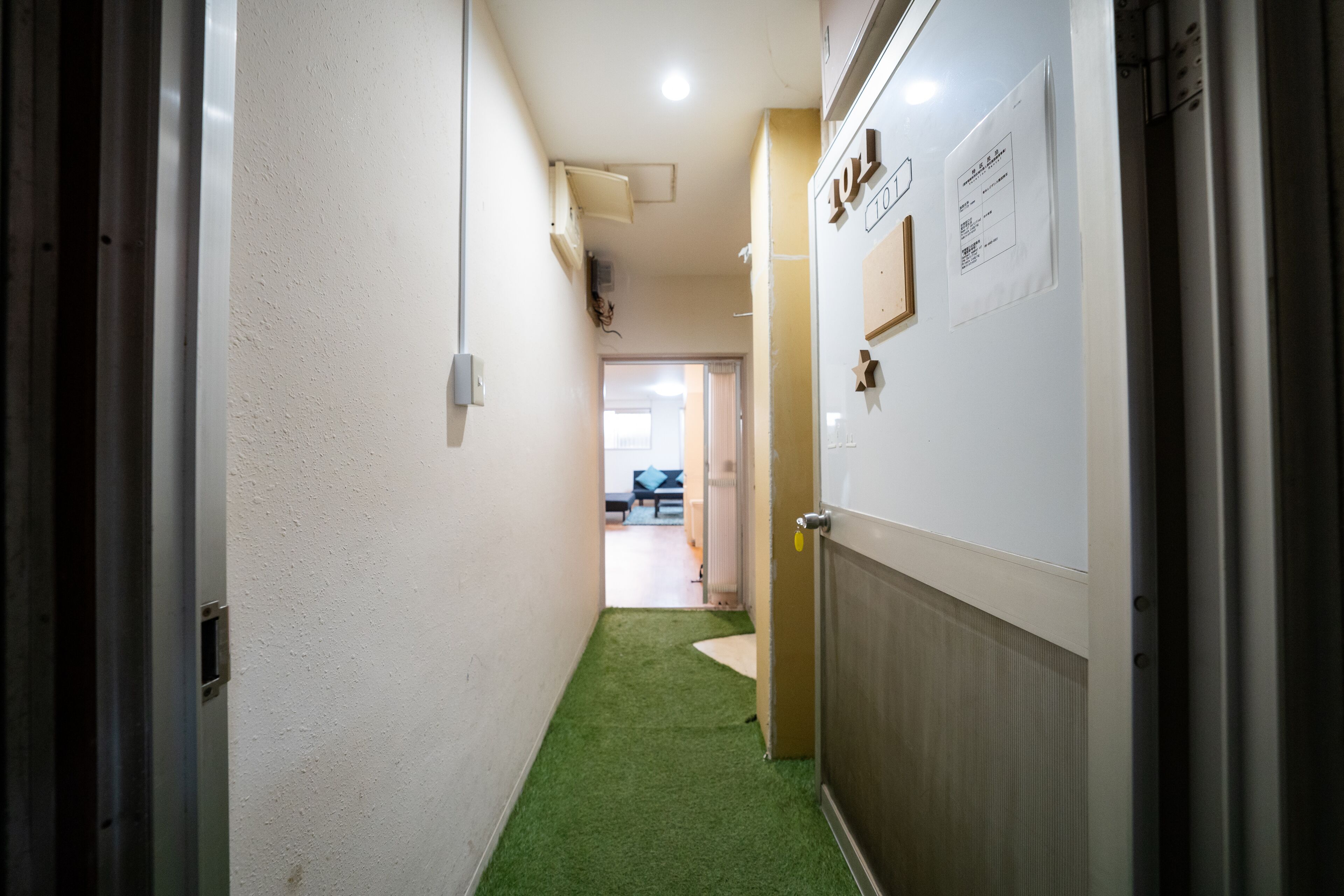 Un couloir avec un tapis vert synthétique, une porte en bois et un mur blanc orné d’un tableau d’affichage.